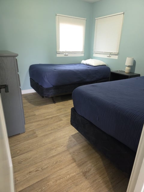 Light blue bedroom with two twin beds with dark blue bedding, wooden floor, and two windows with white blinds