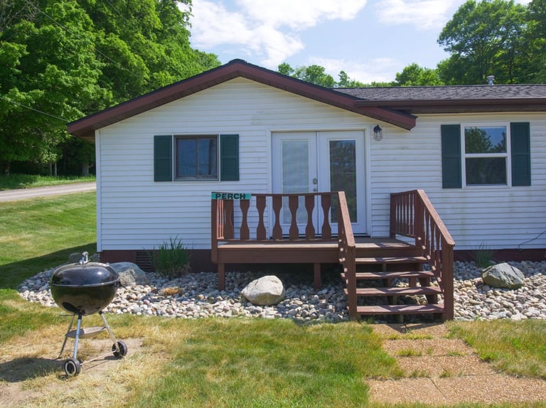 White mobile home with dark brown roof and wooden deck, surrounded by trees, with a grill in the front yard