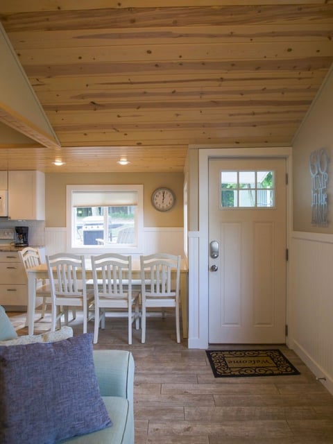 Bright kitchen and dining area with wooden ceiling beams, white chairs at a round table, and white cabinetry overlooking windows