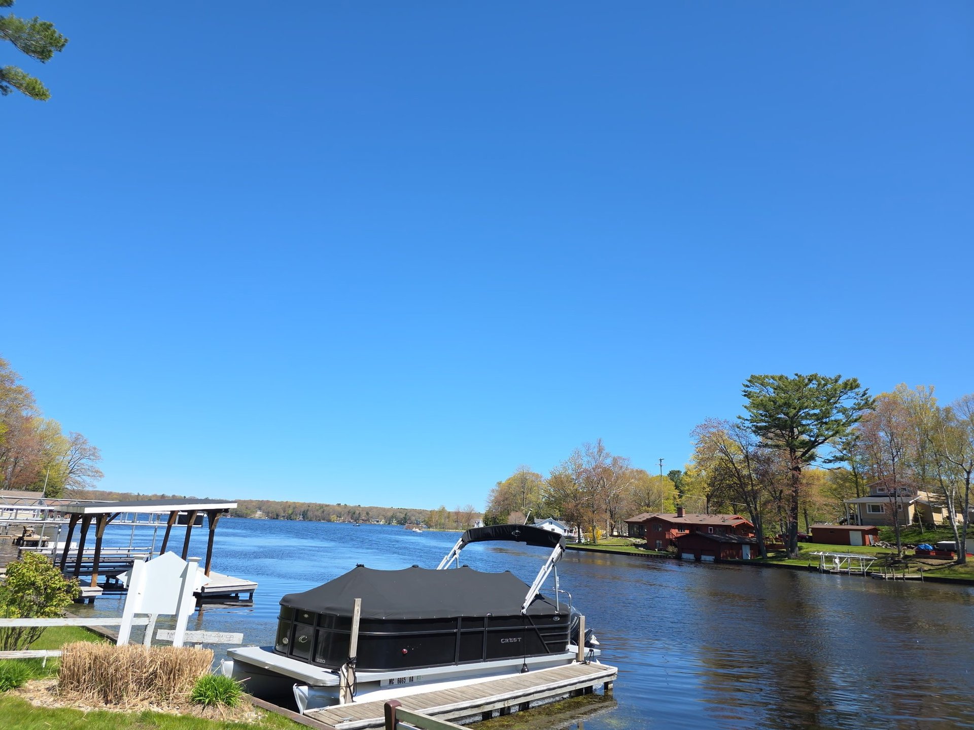 Black pontoon boat docked on calm blue river with trees and docks visible along the shoreline under clear sky