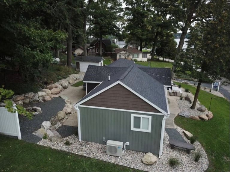 Aerial view of a residential property with a green-and-white cottage, gabled roof, gravel landscaping, and trees surrounding the home