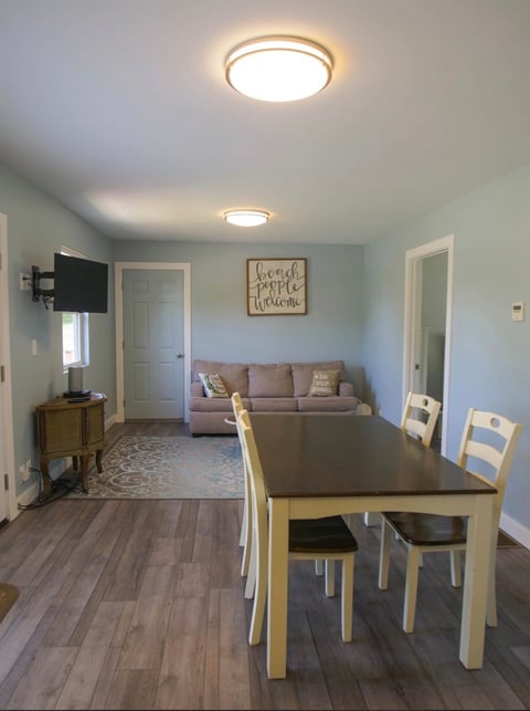 Open-plan living space with dining table, chairs, and beige sofa, light wood flooring, mounted TV, and modern ceiling lights
