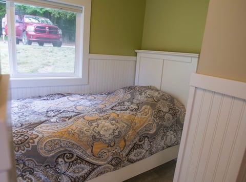 Cozy bedroom with a bed covered in patterned bedding, white wainscoting on walls, olive green accent wall, and window overlooking a parked red truck outside