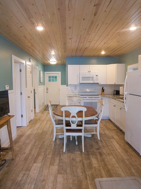 Bright kitchen with turquoise walls, white cabinets, wood ceiling, round dining table with white chairs, and wooden plank flooring