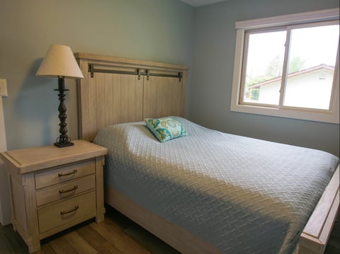Cozy bedroom with a neatly made bed, wooden nightstand with lamp, and large window with natural light