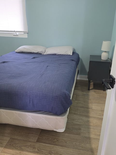 A bedroom with a bed covered in blue bedding and white pillows, a black nightstand with a lamp, and light blue walls