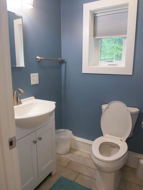 Modern bathroom with blue walls, white vanity, sink, and toilet, featuring a window with blinds and light tile flooring
