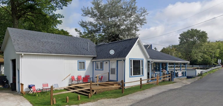 Small white and dark blue cottage-style house with front porch, wooden railings, and colorful patio furniture under trees