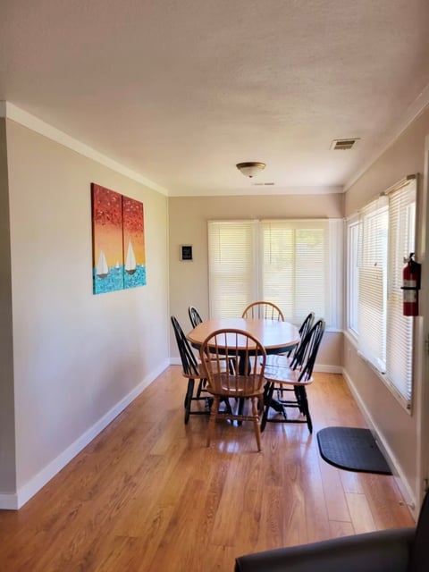 Bright dining room with wooden table and four chairs, artwork on wall, large windows with blinds, and hardwood flooring