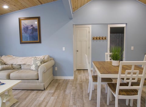 Modern studio apartment with cream sofa on left, dining table with white chairs on right, blue accent wall, and wood plank flooring throughout