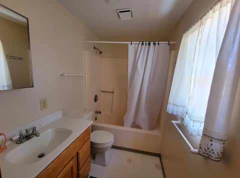 Clean bathroom with white sink, wooden vanity, toilet, shower with curtain, and window with natural light
