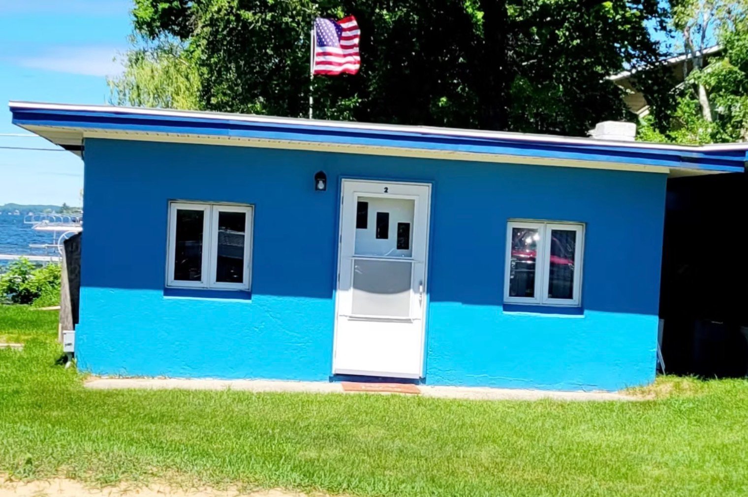 Blue and turquoise small building with white door and windows, American flag on roof, sitting on grass near water
