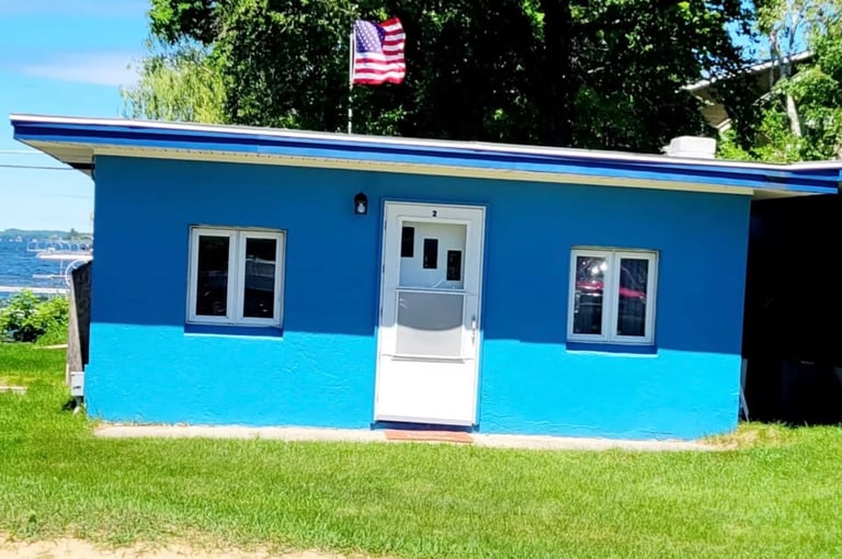 Blue and turquoise small building with white door and windows, American flag on roof, sitting on grass near water