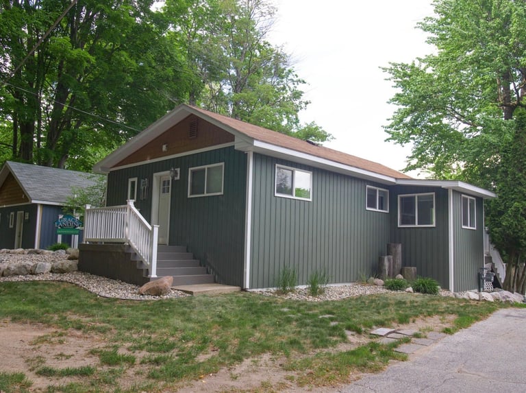 Dark green corrugated metal cabin with brown roof, white trim, and front porch surrounded by trees and gravel grounds