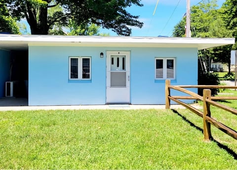Light blue single-story residential building with white door and windows, surrounded by green lawn and wooden fence