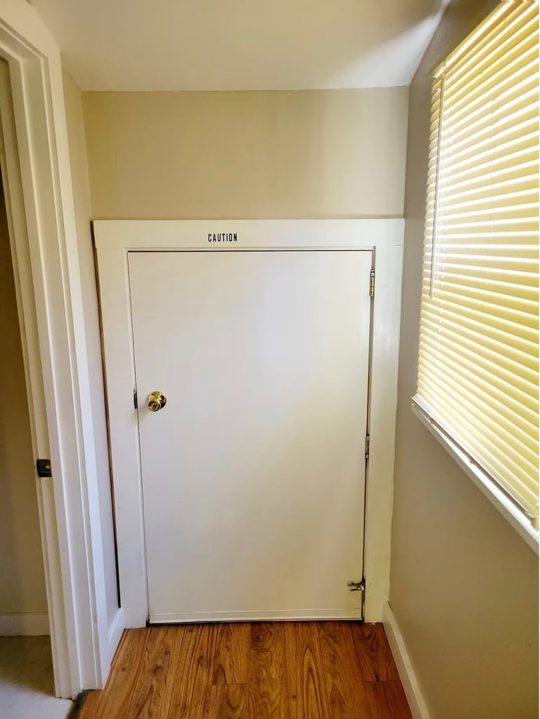 Interior hallway with white caution-labeled electrical panel door, wood flooring, cream walls, and blinds on window