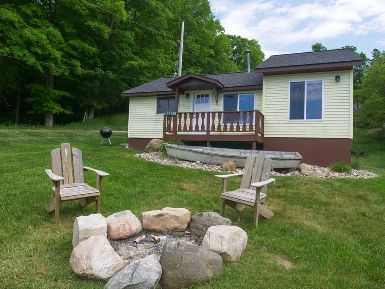 Small cottage with wooden deck surrounded by green trees, two Adirondack chairs facing a fire pit with stones in grassy yard