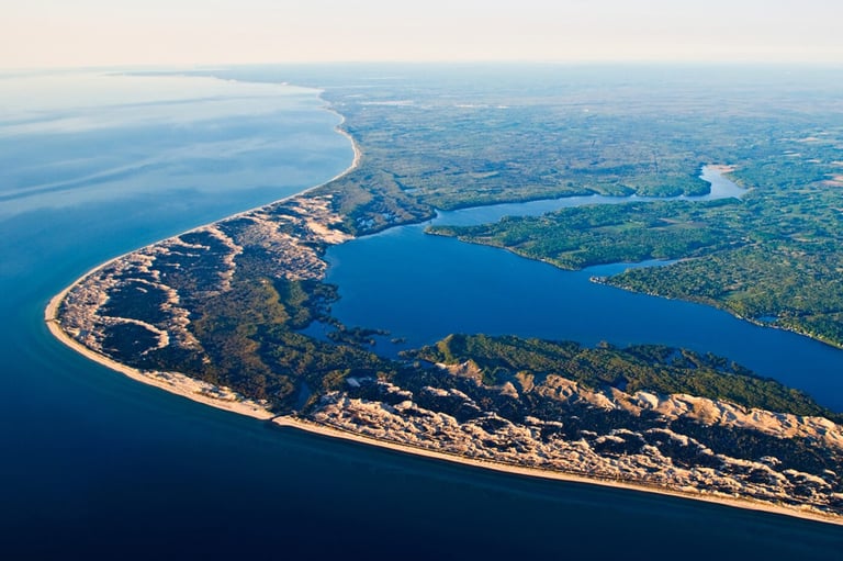 Aerial view of a curved coastline with sandy peninsula, blue water, and forested inland areas