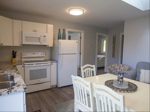 Modern kitchen and dining area with white cabinets, stainless steel appliances, white dining table, and gray seating visible in background