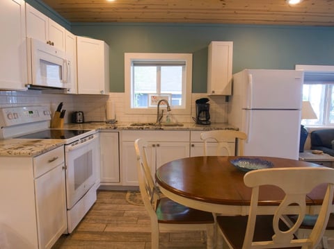 Bright kitchen with cream cabinetry, white appliances, granite countertops, and wooden dining table with chairs in foreground