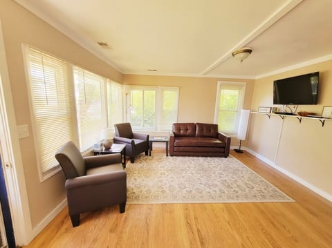Bright, spacious living room with cream walls, hardwood floors, brown leather furniture, large windows with natural light, and a wall-mounted TV