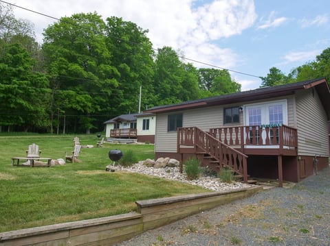 Rural home with wooden deck surrounded by green lawn, fire pit, and mature trees under blue sky