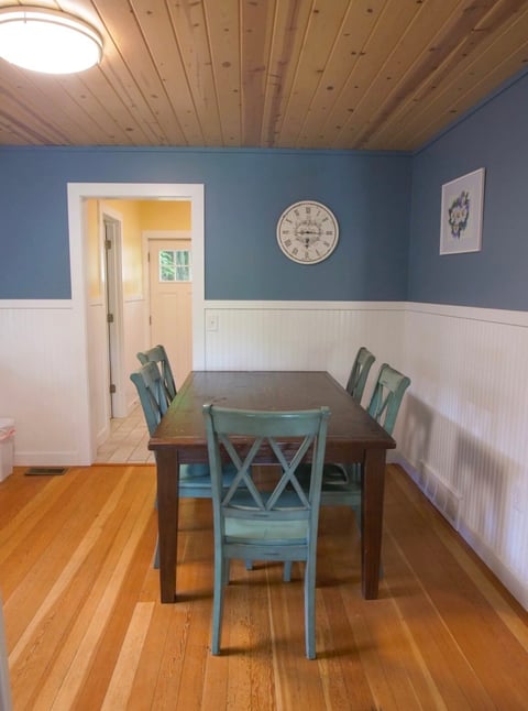 Dining room with wood table, two turquoise chairs, blue walls with white wainscoting, and wooden ceiling with round clock