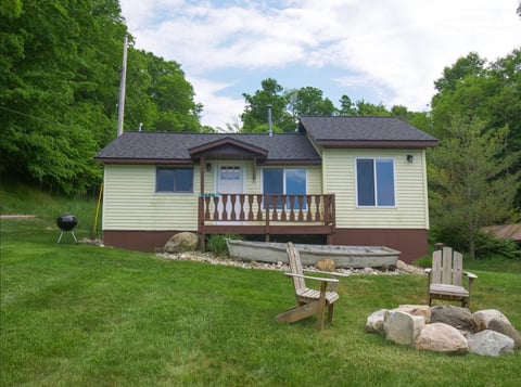 Single-story beige cottage with dark gray roof, wooden deck, and surrounded by green lawn and trees