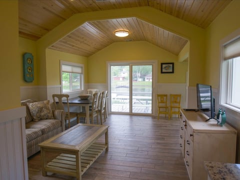 Bright yellow cottage interior with vaulted ceiling, wood flooring, seating area, dining table, and glass doors overlooking exterior