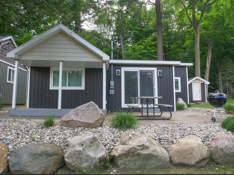 Modern dark gray cottage with white trim on gravel ground, surrounded by trees and large boulders at waterfront