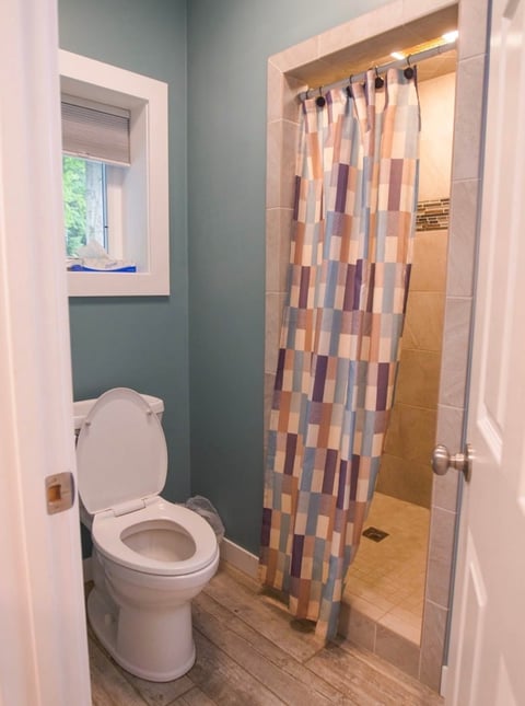 Bathroom interior with white toilet on left, shower with geometric patterned curtain on right, light blue-gray walls, and small window above toilet