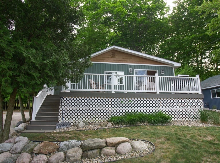 Elevated residential home with white deck railing and lattice skirting surrounded by green trees and landscaping rocks