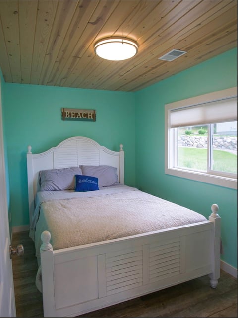 Bright turquoise bedroom with white wooden bed, wooden ceiling, and window overlooking green landscape