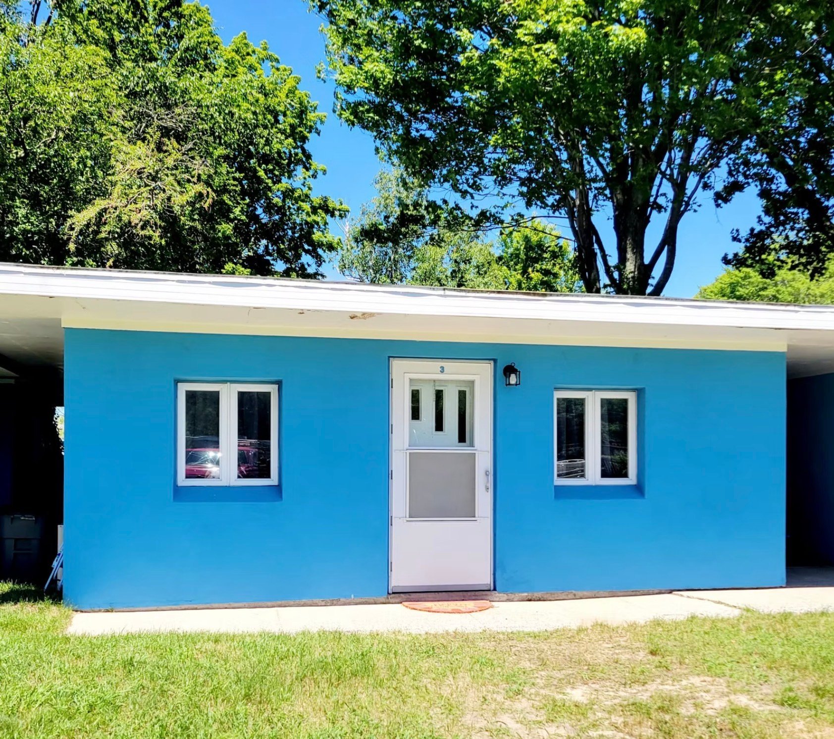 Bright blue modern house with white door and windows, surrounded by green lawn and large trees under clear blue sky