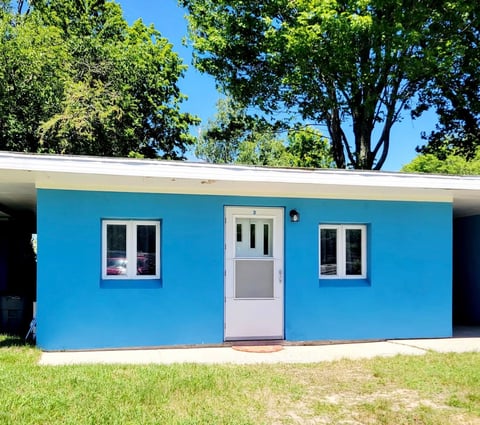 Bright blue modern house with white door and windows, surrounded by green lawn and large trees under clear blue sky